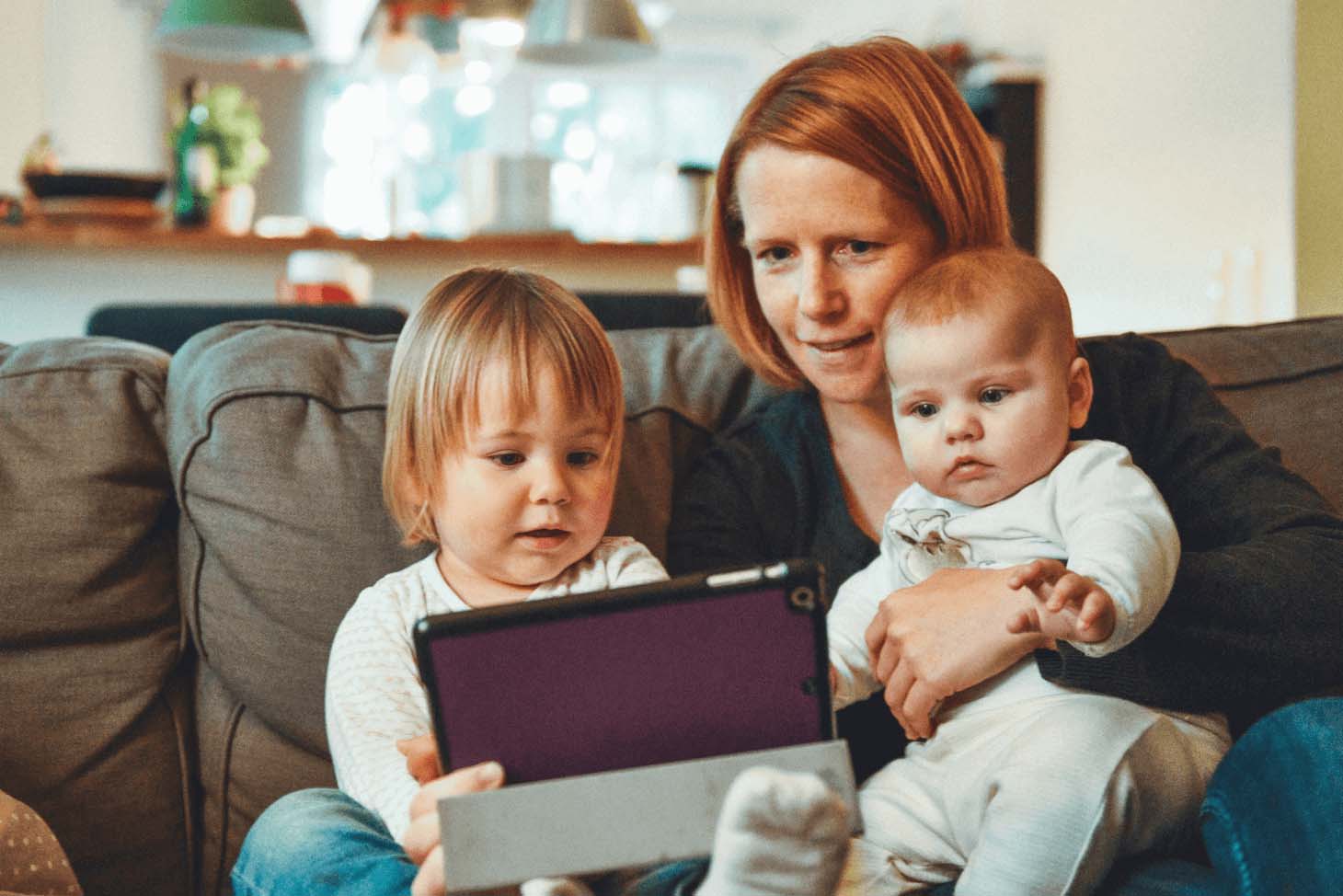A woman accompanied by two children on a sofa while looking at a tablet.